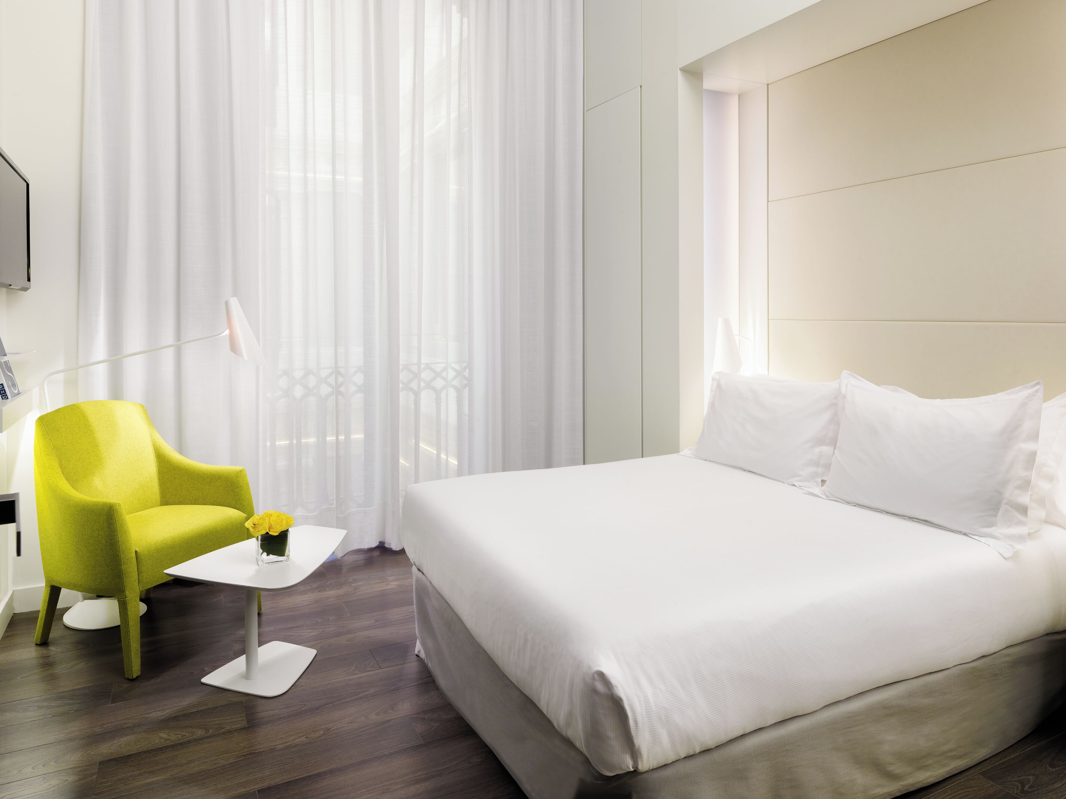 Una habitación de hotel con una cama, un sillón amarillo brillante, una mesita blanca pequeña y cortinas de gasa.