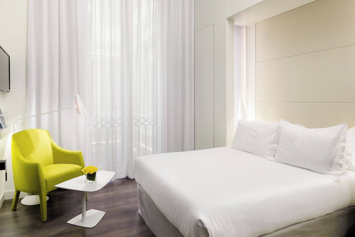 Ein Hotelzimmer mit einem großen Bett, einem leuchtend gelben Sessel, einem kleinen weißen Tisch und transparenten Vorhängen.