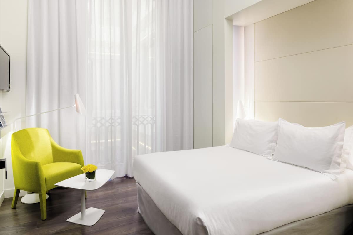 Una habitación de hotel con una cama, un sillón amarillo brillante, una mesita blanca pequeña y cortinas de gasa.