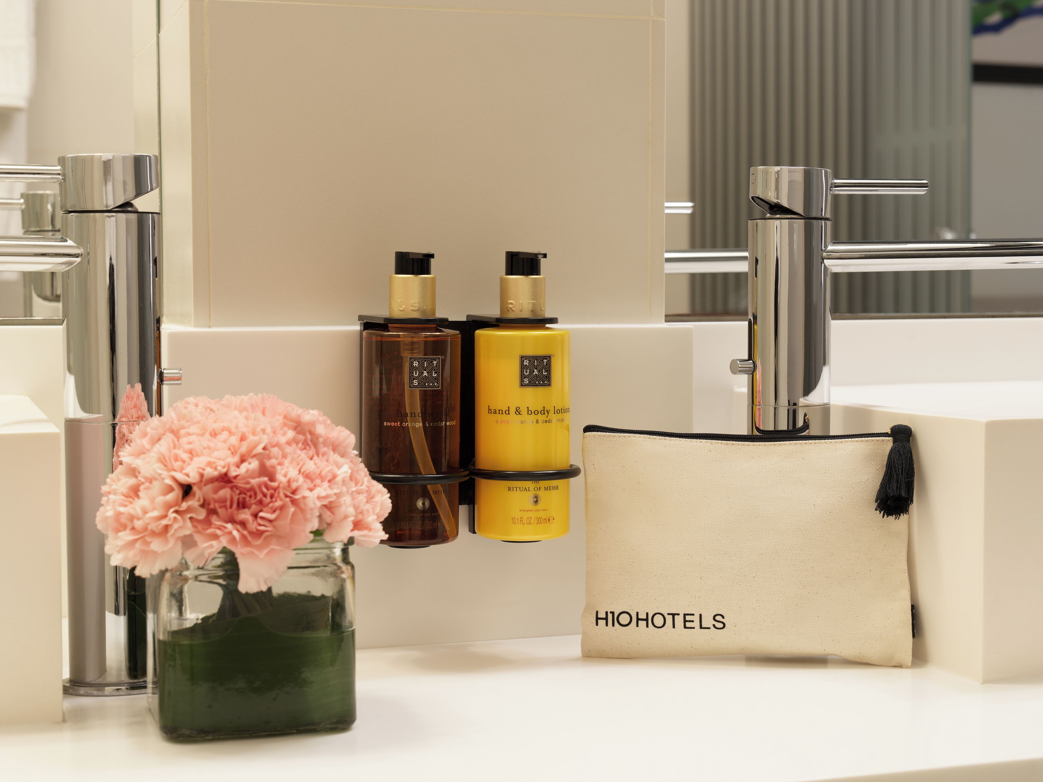 A bathroom with a mirror, two bottles of lotion, a vase with pink flowers, and a toiletry bag with 'HIOHOTELS' text.