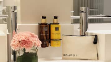 A bathroom with a mirror, two bottles of lotion, a vase with pink flowers, and a toiletry bag with 'HIOHOTELS' text.