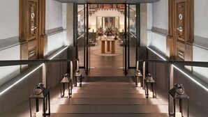 A grand hallway with a high arched glass door leading to a lobby area, flanked by wooden doors and illuminated by floor lamps.