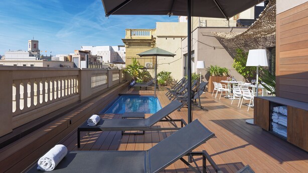 A rooftop pool area with lounge chairs, a table with chairs, and a bar area with a wooden backdrop.