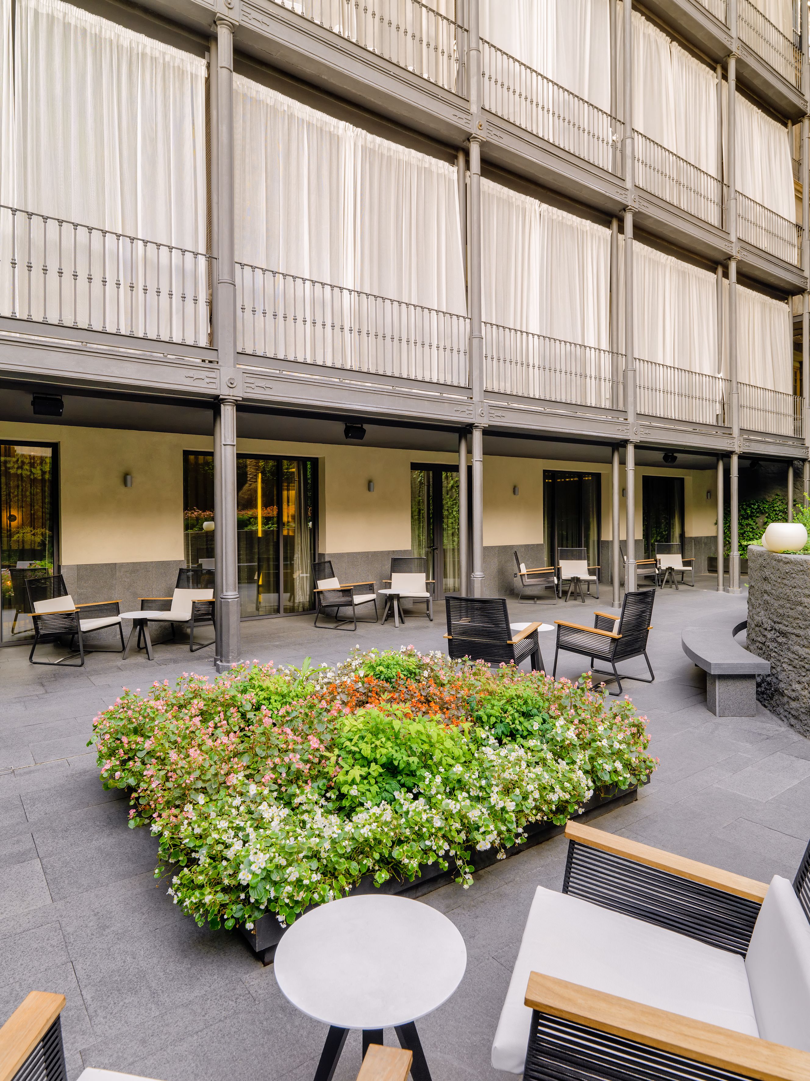 Un patio con mesas al aire libre, un parterre de flores y un edificio con balcones.