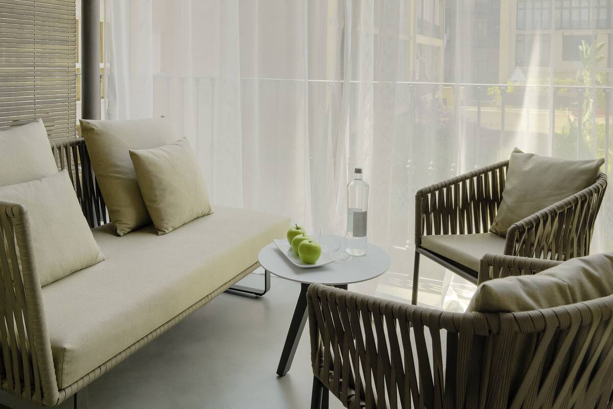 Ein modernes Wohnzimmer mit einer Couch, zwei Sesseln, einem kleinen Tisch mit einer Flasche und Äpfeln sowie durchsichtigen Vorhängen.