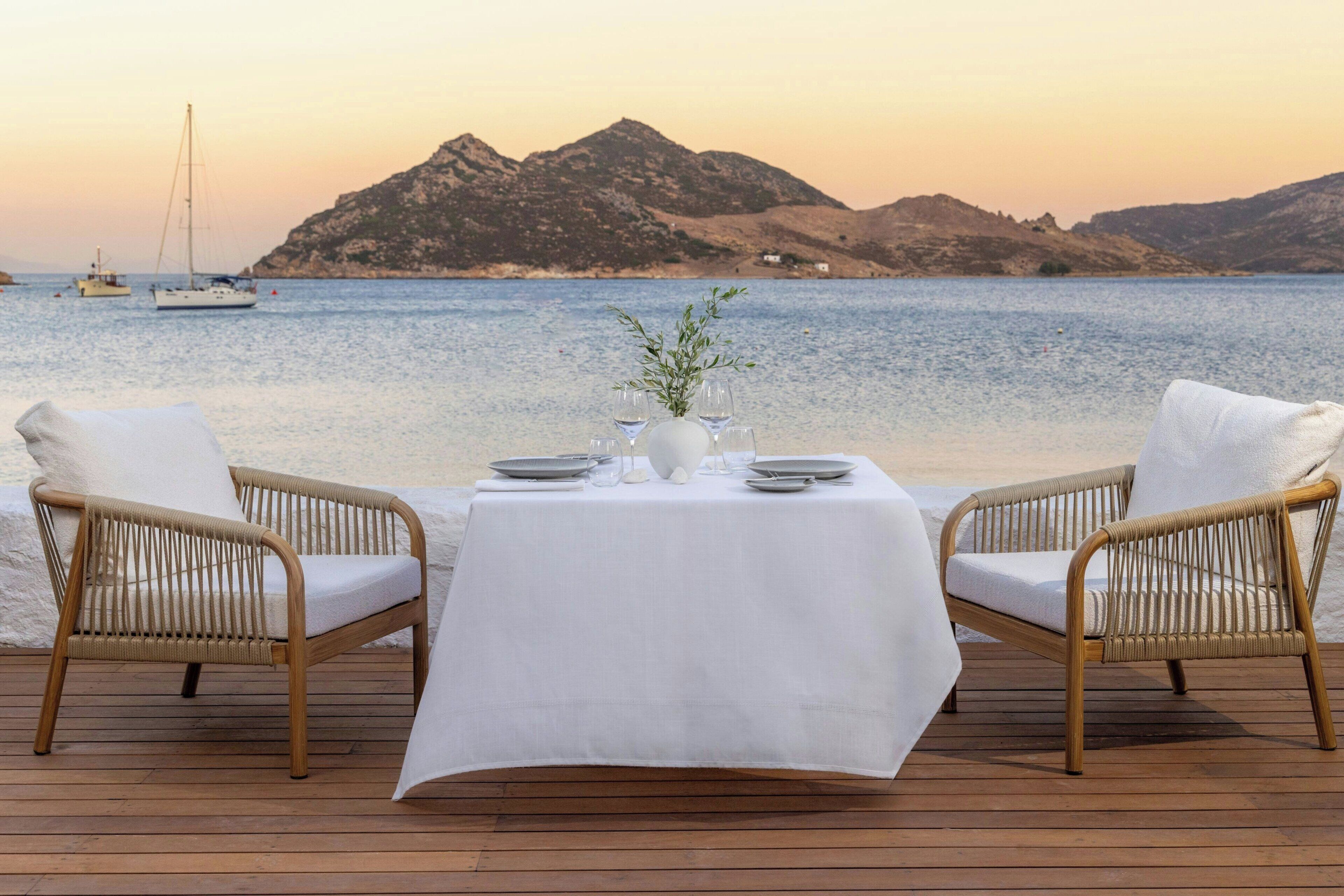 A dining table set on a wooden deck with wicker chairs, overlooking a calm sea and distant hills.