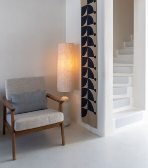 A chair with a light grey fabric and a patterned pillow, a tall floor lamp with a textured shade, and a white staircase with a black geometric wall panel. - Patmos Aktis, a Luxury Collection Resort & Spa, Greece (Patmos)
