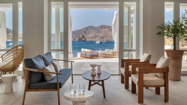 A spacious room with a large window overlooking a scenic coastal view, furnished with a sofa, chairs, and a small table. - Patmos Aktis, a Luxury Collection Resort & Spa, Greece (Patmos)