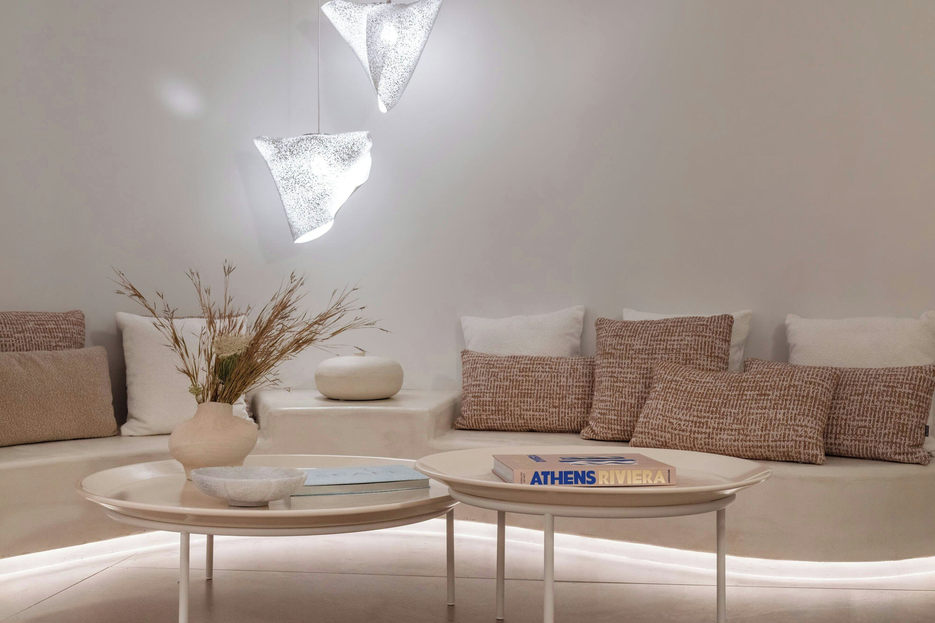Una sala de estar moderna con un sofá, dos mesas redondas, un jarrón con plantas secas y un libro titulado 'ATHENS RIVIERA'.