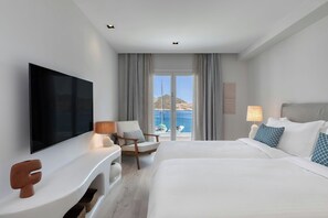 A hotel room with a large bed, a TV, a desk, and a chair. There is a window with a view of the sea and a mountain. - Patmos Aktis, a Luxury Collection Resort & Spa, Greece (Patmos)