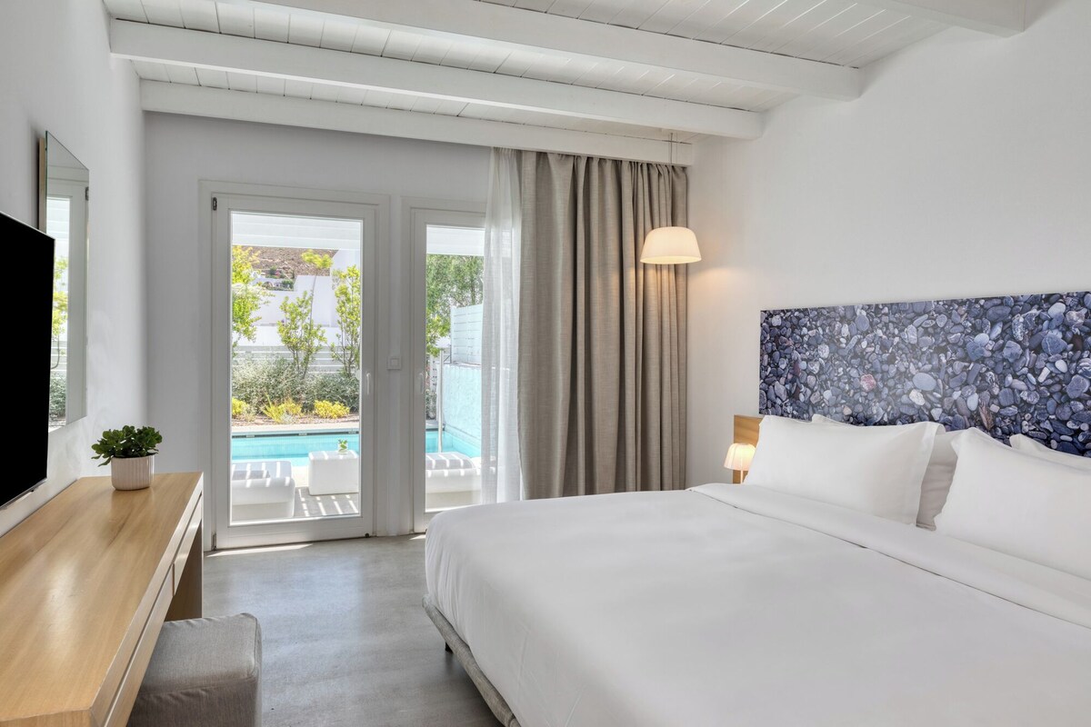 Habitación de hotel con cama, un escritorio con una planta, televisor, ventana grande con vista a una piscina y vegetación, y techo con vigas a la vista.