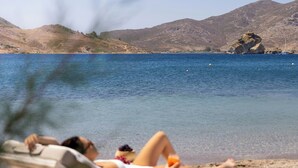 A beach scene with a person reclining on a sun lounger, a clear view of the sea, and a mountainous backdrop. - Patmos Aktis, a Luxury Collection Resort & Spa, Greece (Patmos)