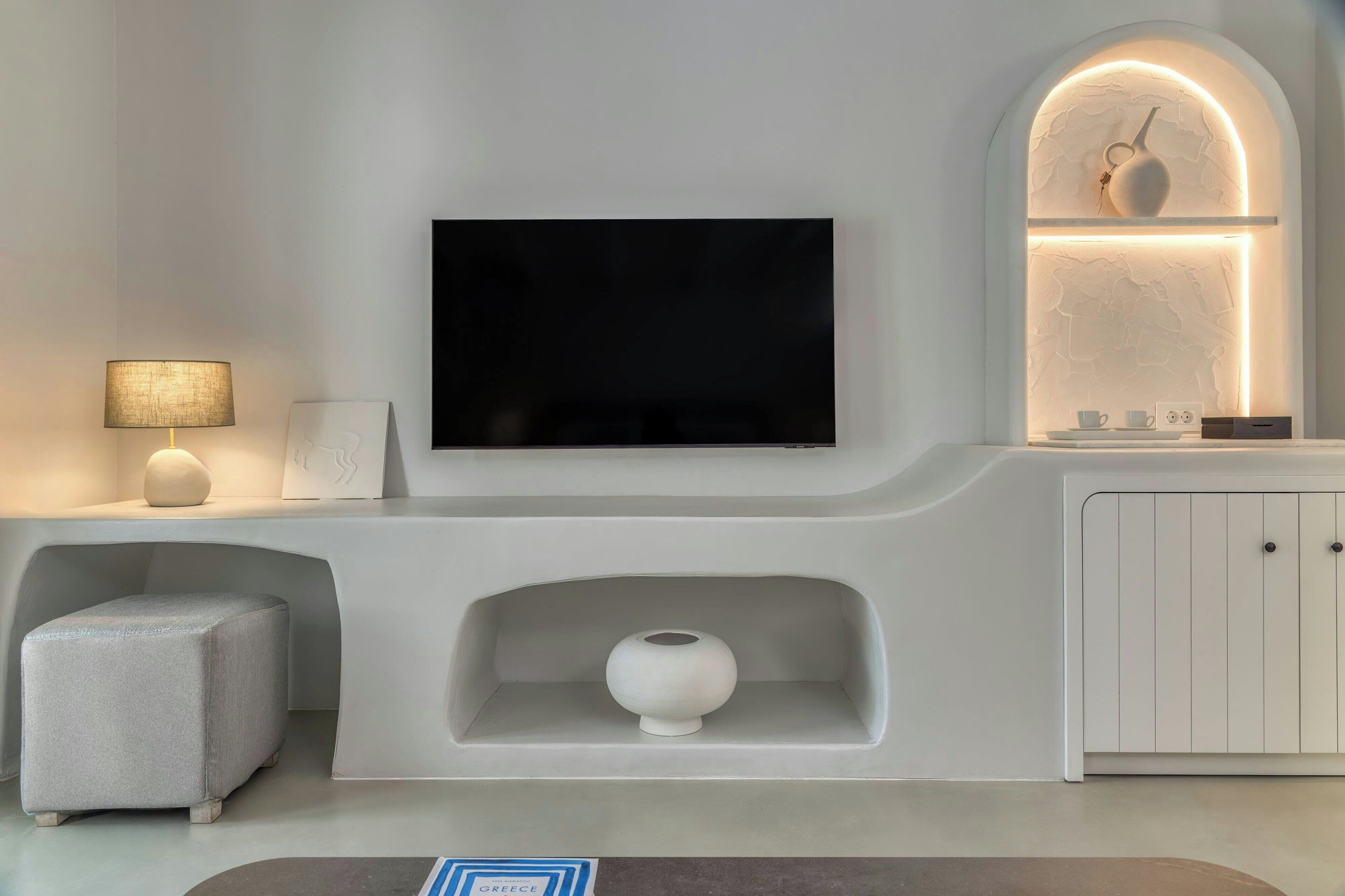 A modern living room with a flat-screen TV mounted on a white wall, a small white cabinet below, and a grey ottoman to the left.