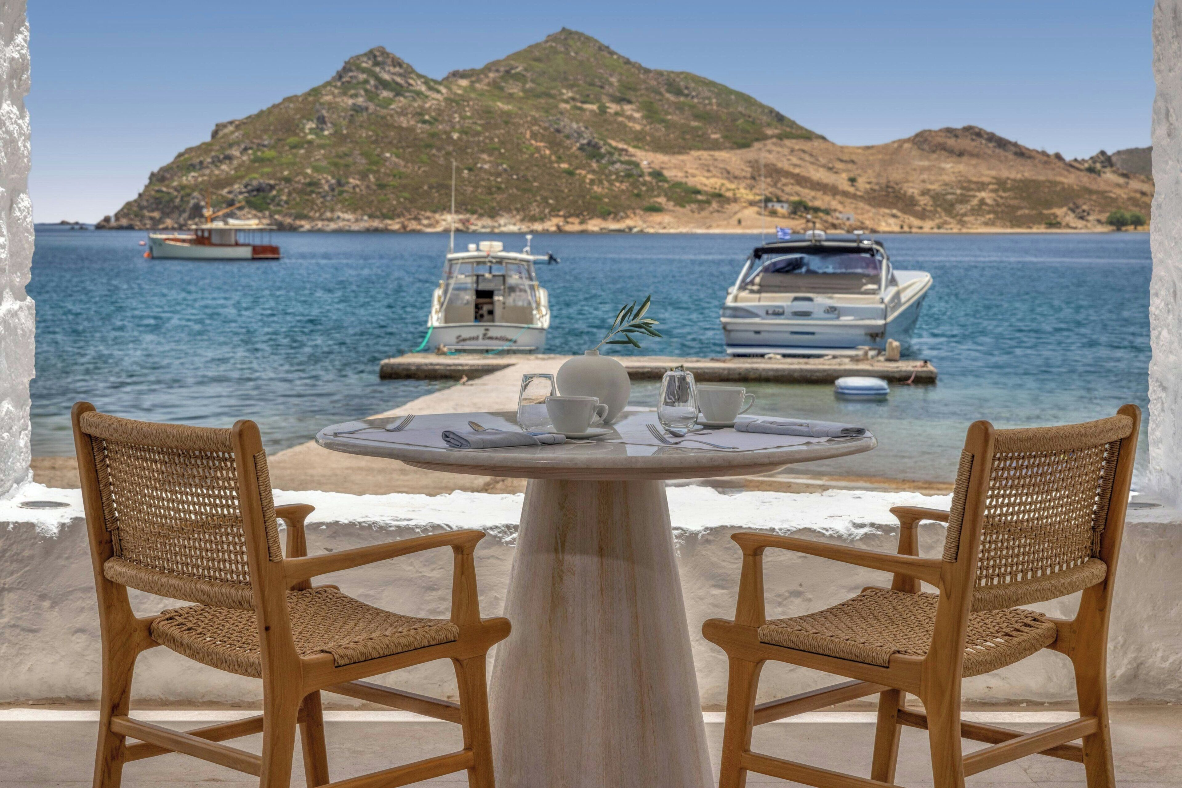 Uma mesa com duas cadeiras de vime voltada para um corpo d'água, com barcos e uma montanha ao fundo.