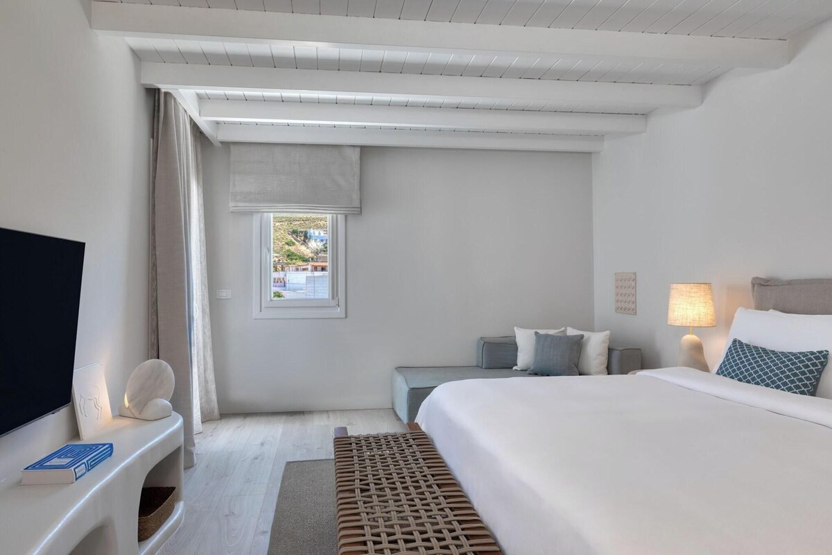 Una habitación de hotel moderna con una cama grande, televisión, un pequeño rincón de descanso y una ventana con cortinas.