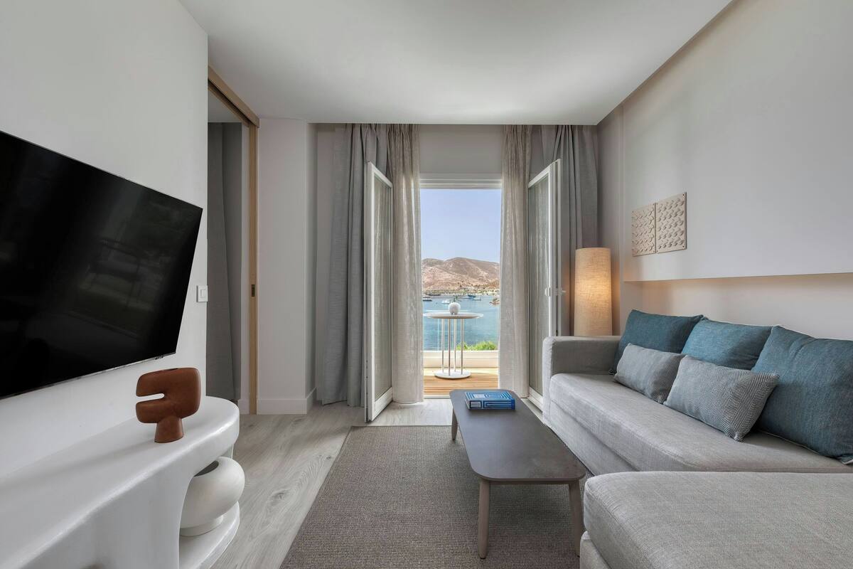 Ein modernes Hotelzimmer mit Flachbildfernseher, einer Couch mit gemusterten Kissen, einem Couchtisch und Blick auf eine gebirgige Landschaft durch große Fenster.