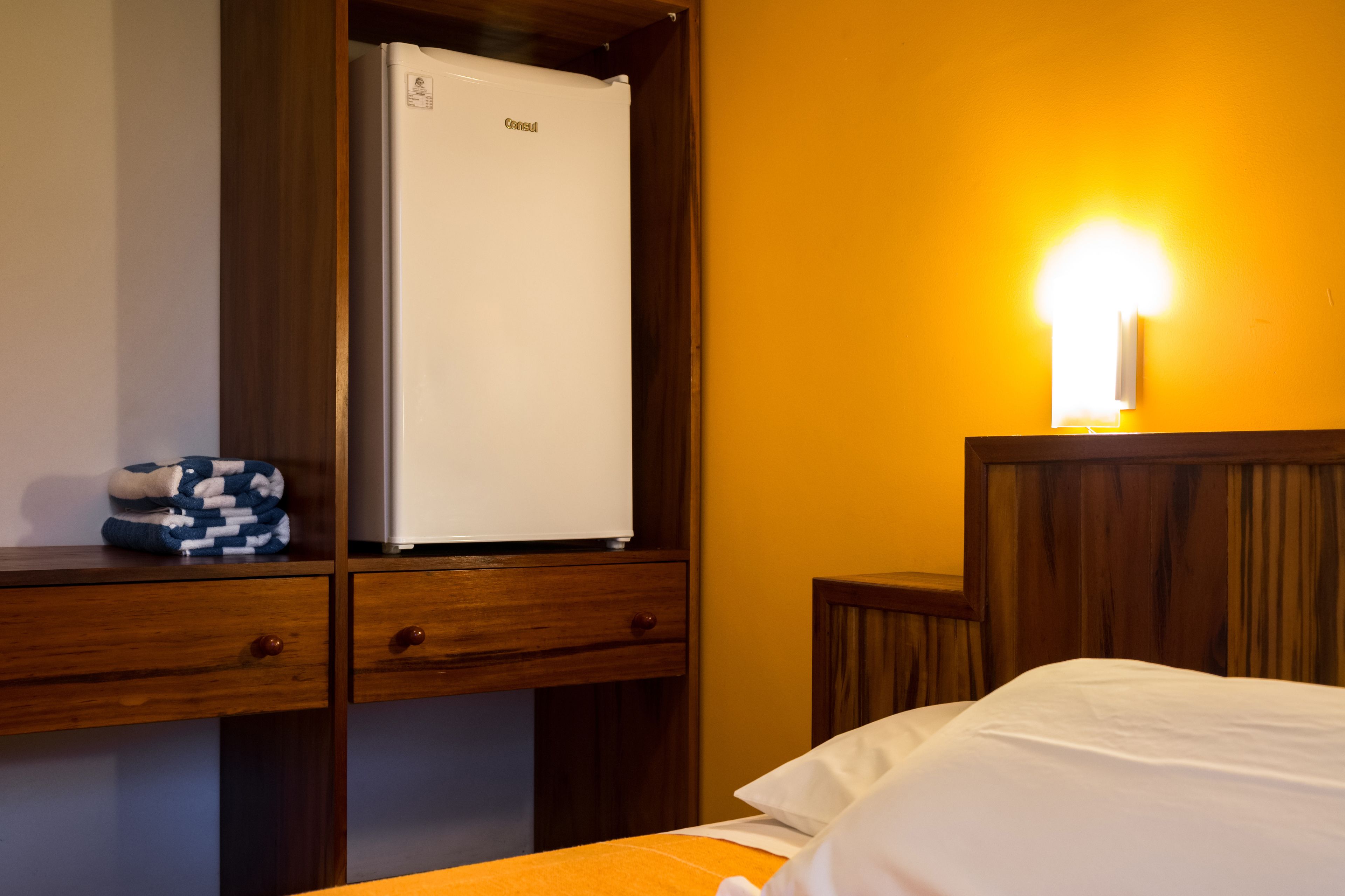 A hotel room with a bed, a wooden nightstand, a white refrigerator, and a wall-mounted light.