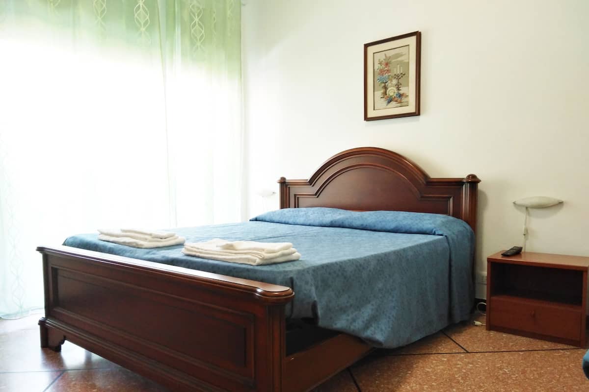A neatly made bed with a blue bedspread and white towels, a wooden headboard, a bedside table with a lamp, and a framed picture on the wall.