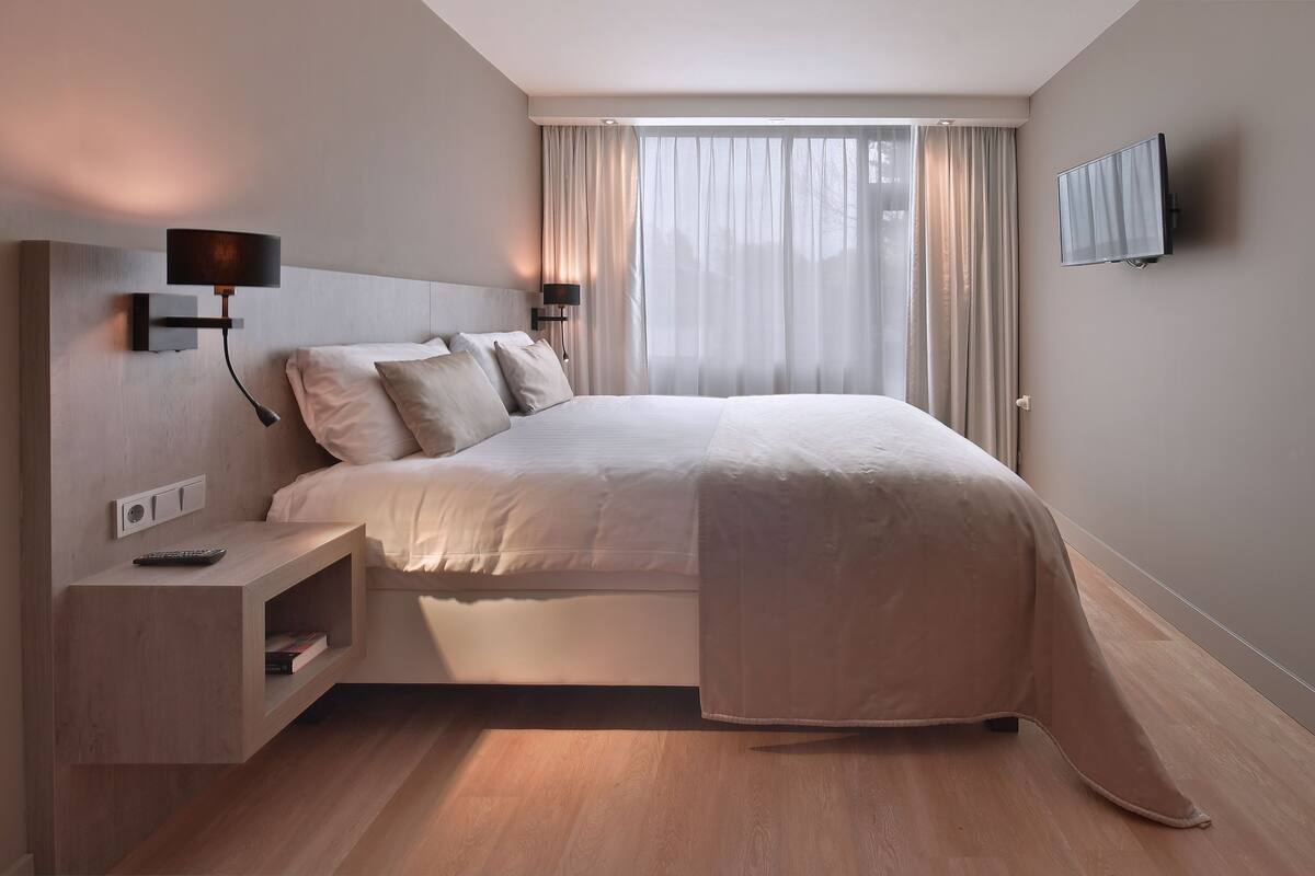 Un dormitorio moderno con cama, mesitas de noche, televisor y ventana con cortinas.