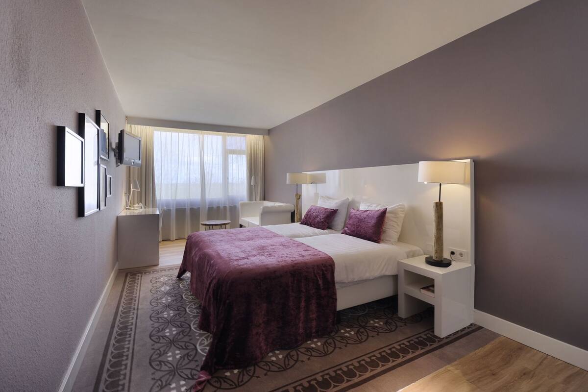 Una habitación de hotel con cama, mesitas de noche, escritorio, silla, ventana con cortinas y alfombra estampada.