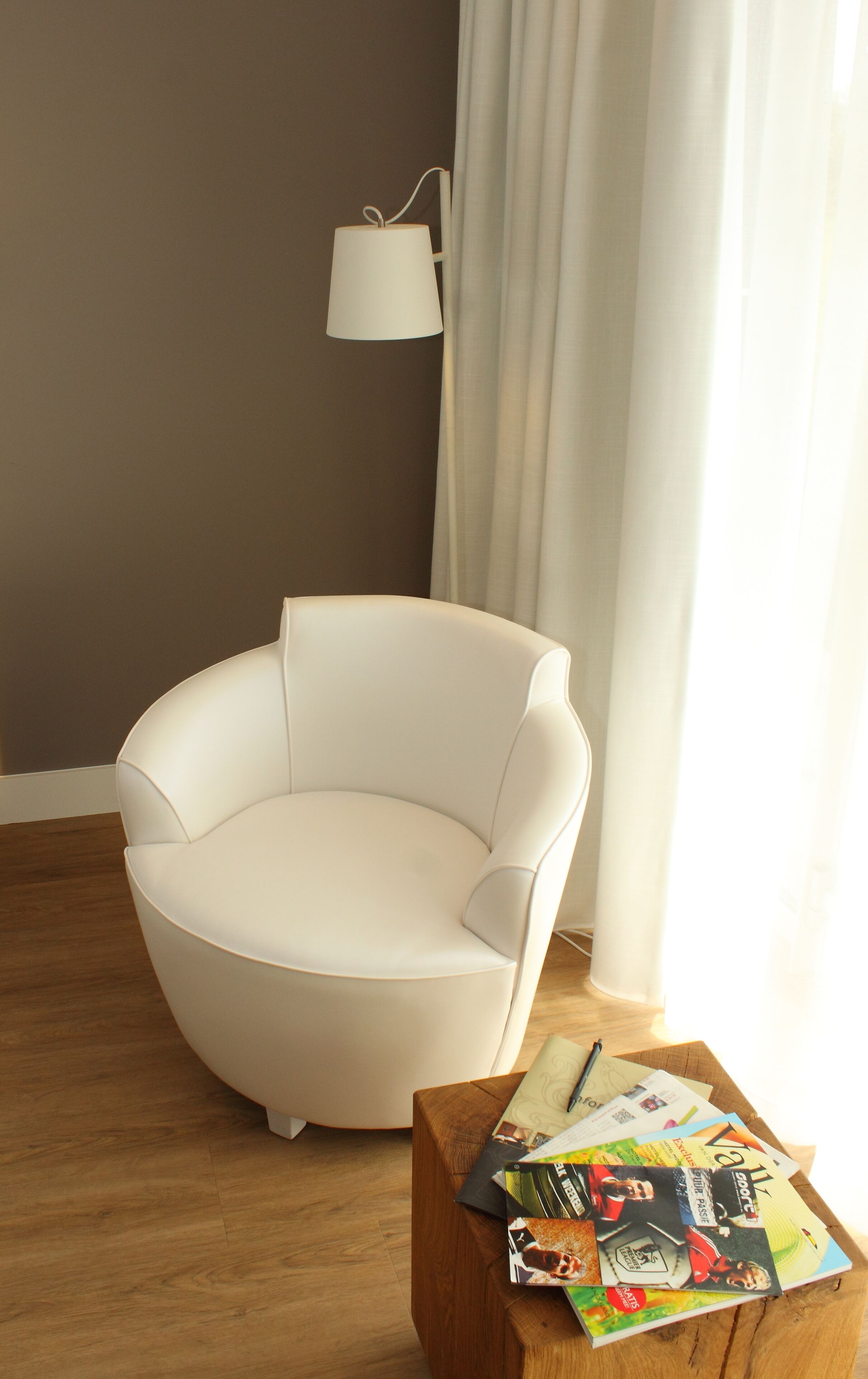 Un salon moderne avec un fauteuil blanc, une table basse en bois sur laquelle sont posés des magazines, un lampadaire blanc et une fenêtre avec des rideaux.