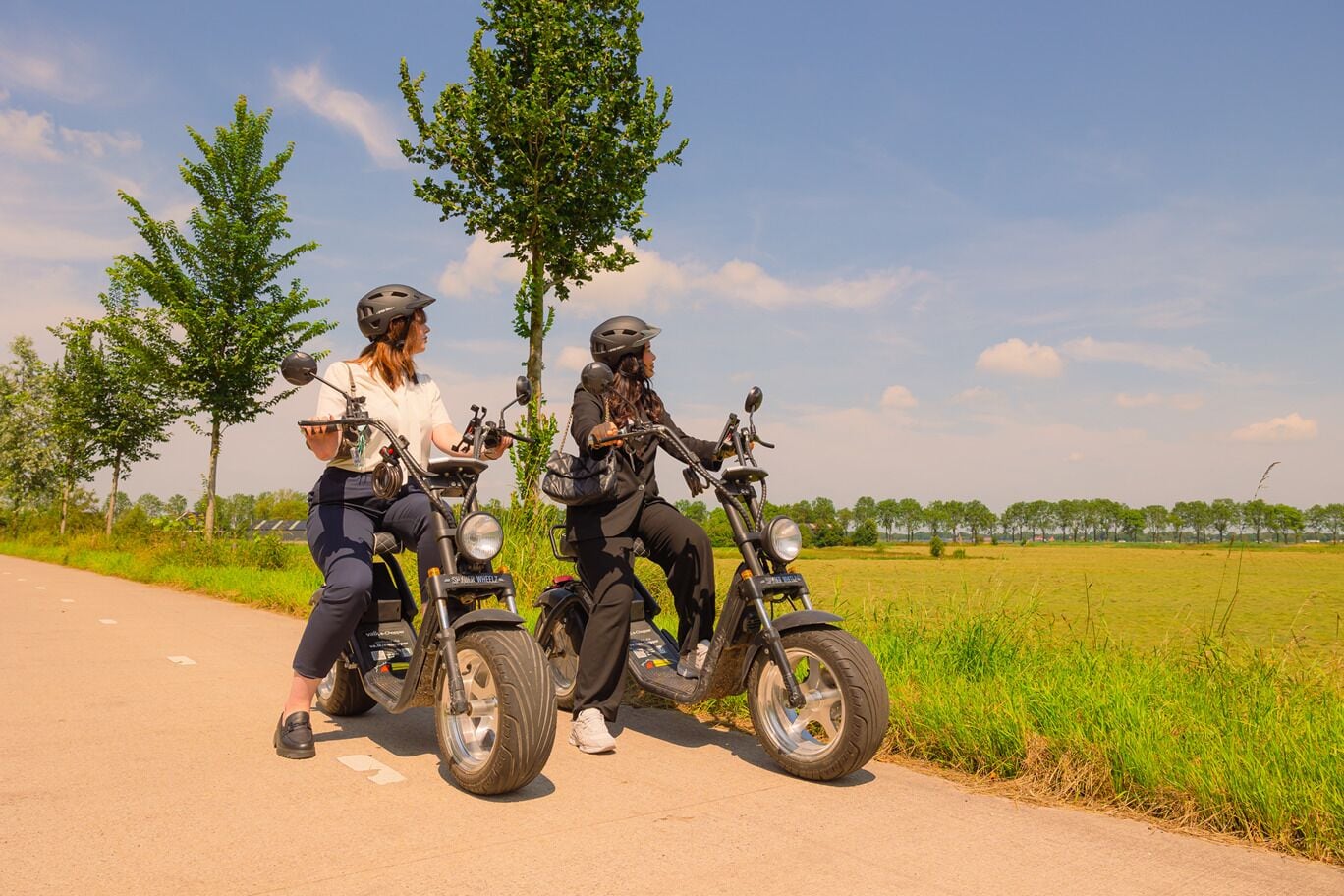 Deux personnes sur une moto, sur un chemin goudronné, avec des arbres et des champs en arrière-plan.
