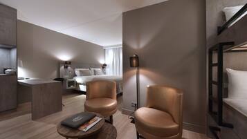A modern hotel room with a bunk bed, a desk, two leather chairs, a bedside table, and a lamp.