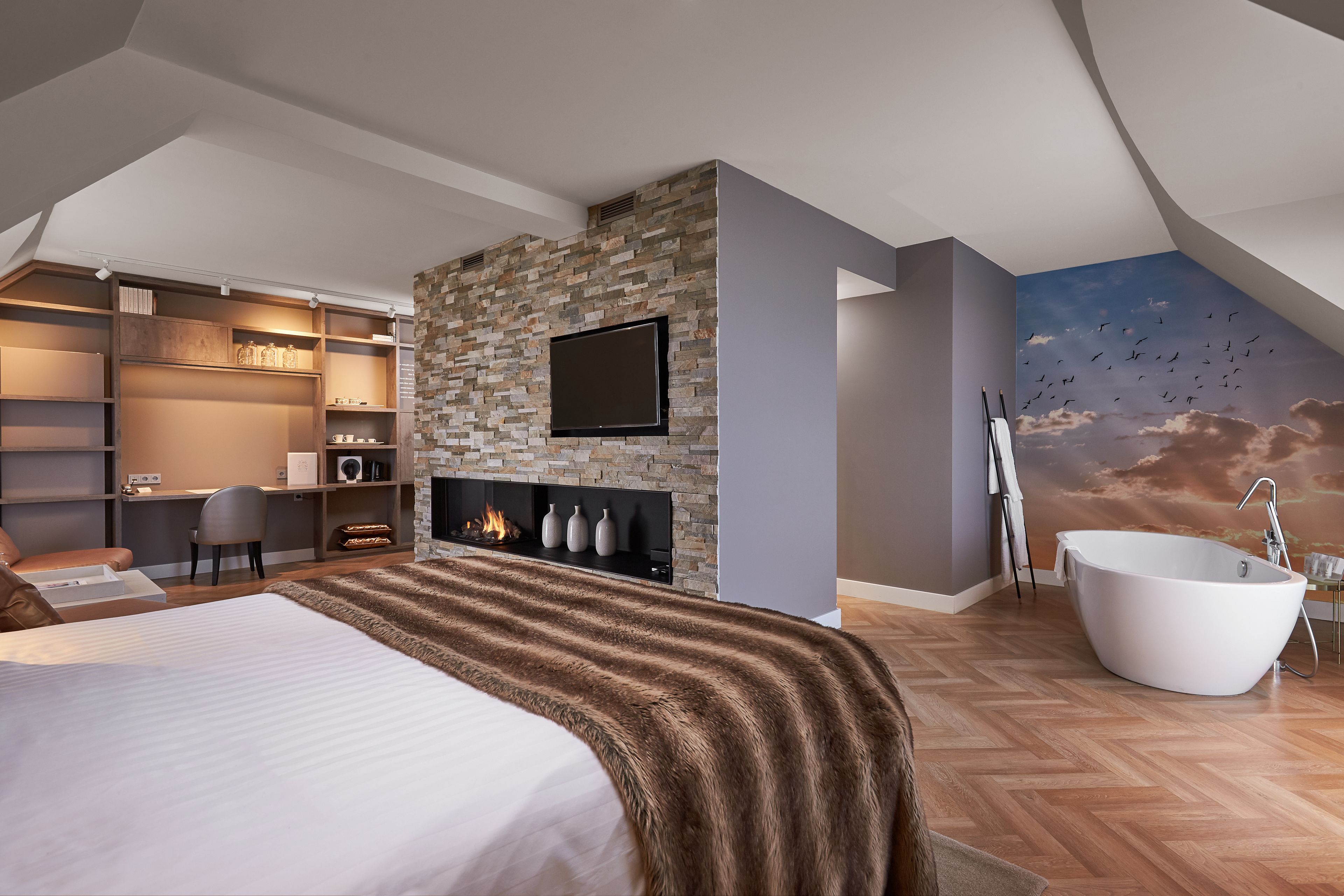 A modern bedroom with a large bed, a fireplace, a TV, a bathtub, and a mural of birds flying.