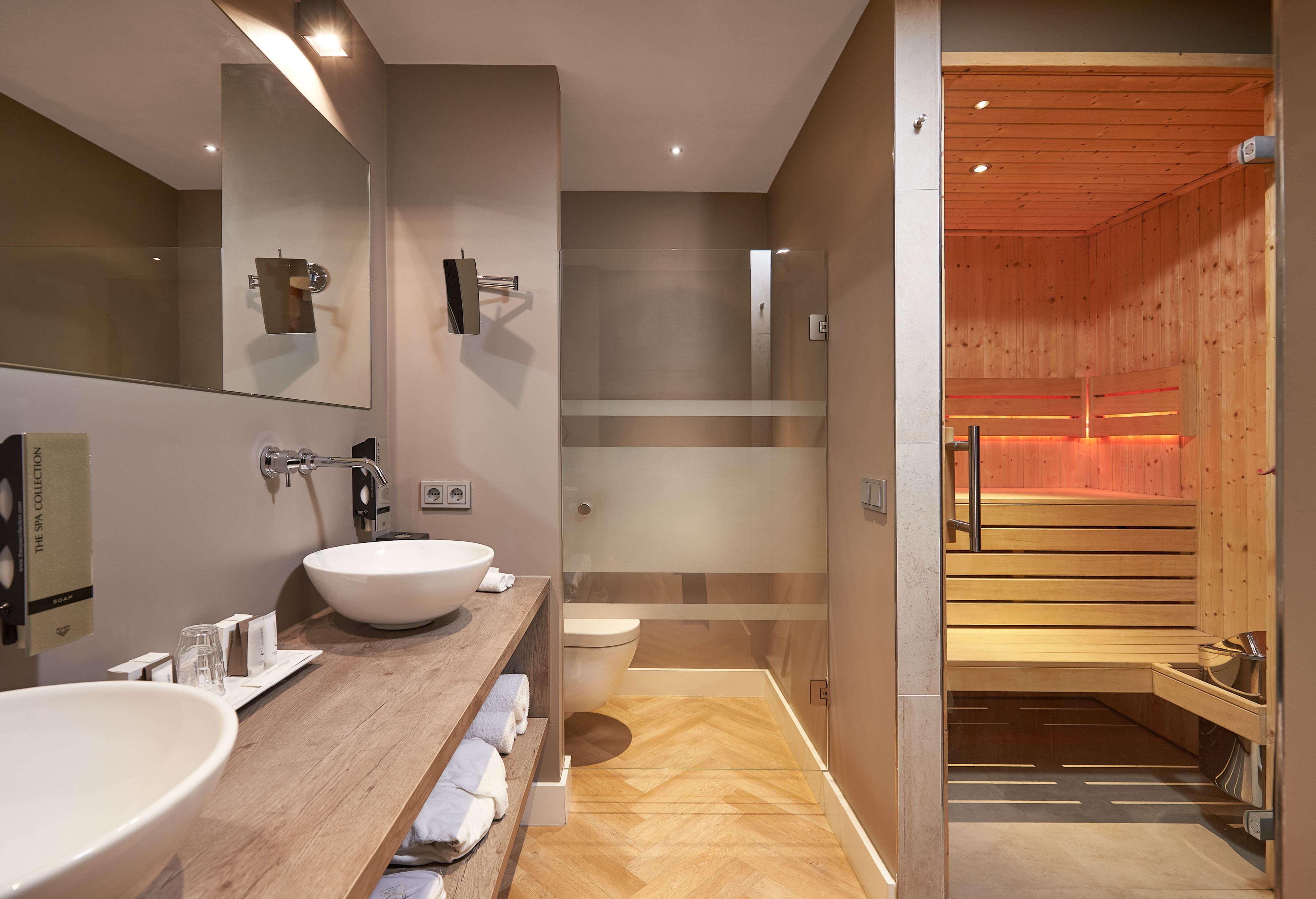 A modern bathroom with a large mirror, two sinks, and a sauna visible through a glass door.