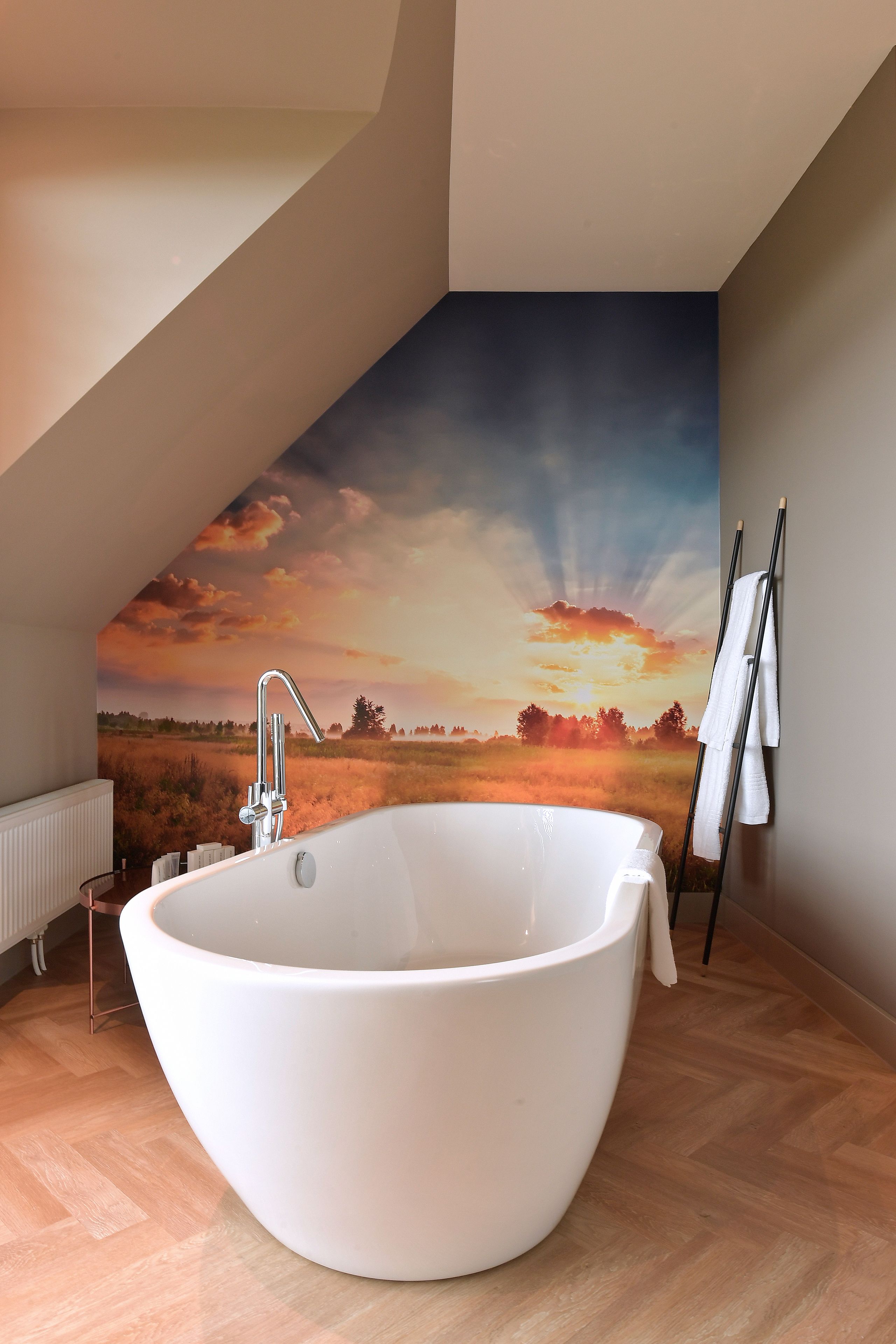 Un baño moderno con una bañera independiente, un radiador y un mural grande que representa un atardecer sobre un campo.