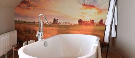 Un baño moderno con una bañera independiente, un radiador y un mural grande que representa un atardecer sobre un campo.