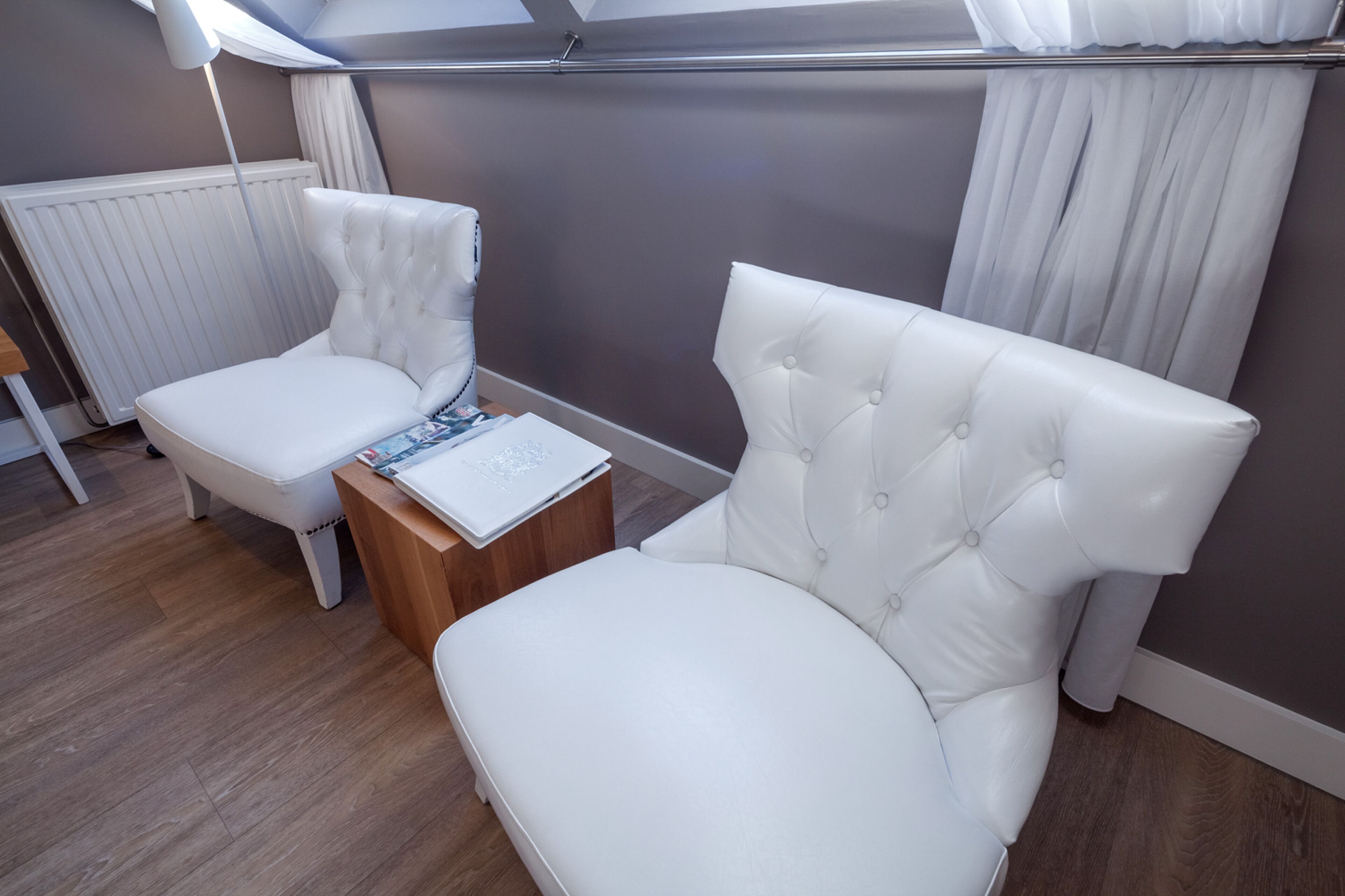 A modern bedroom with a white upholstered bed, a white armchair, and a wooden nightstand with a laptop.