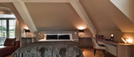 Une chambre avec un grand lit, un bureau et une chaise. Il y a une fenêtre avec des stores et une vue sur la verdure à l’extérieur.