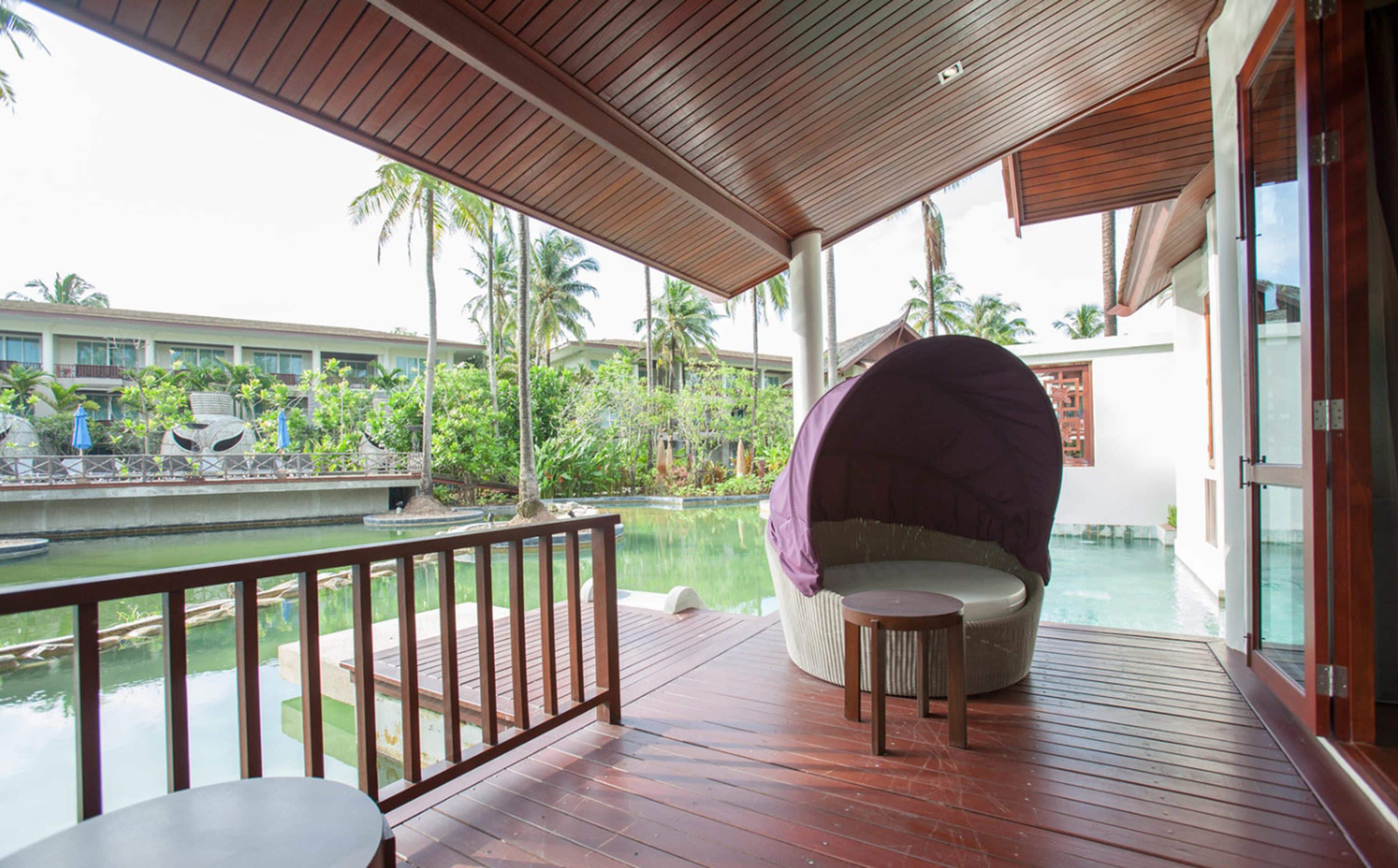 Terraza de madera con zona de estar acolchada con vistas a una piscina y abundante vegetación.