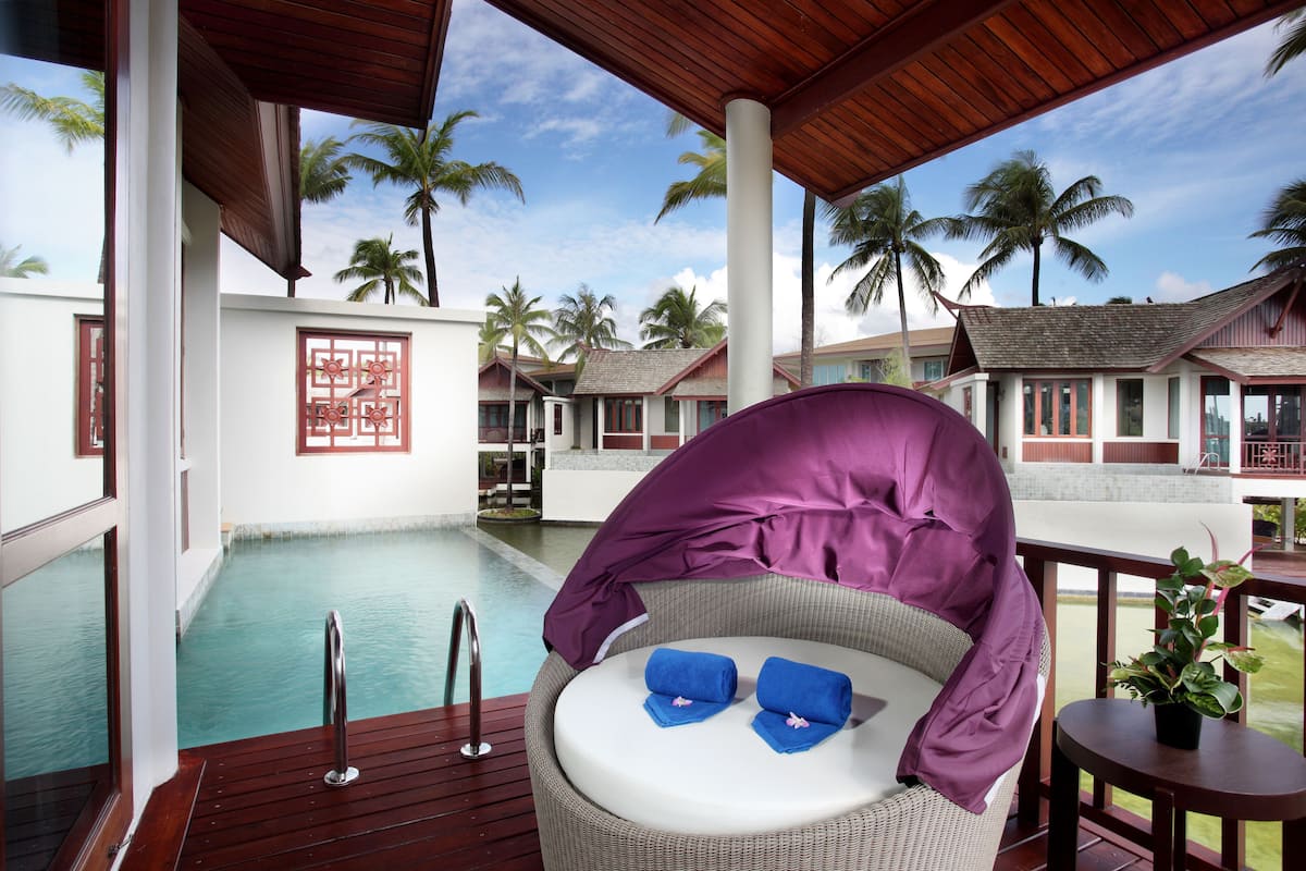 A covered outdoor seating area with a pool, wooden deck, and views of resort buildings and palm trees.