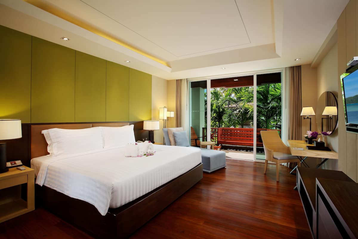 A modern hotel room with a large bed, a desk with a chair, a TV, and a balcony with a view of greenery.