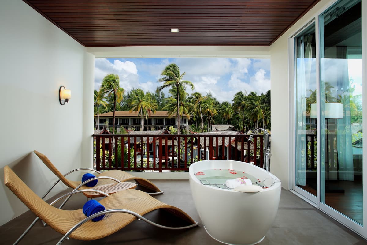 A balcony with two lounge chairs, a white bathtub, and a view of palm trees and a building.