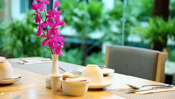 A dining table set with white dishes, a vase with pink flowers, and a view of greenery outside.