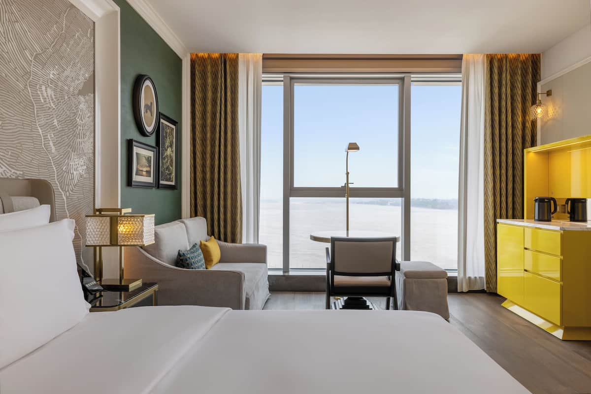 A hotel room with a large window, a bed, a sofa, a small table, and a yellow minibar.