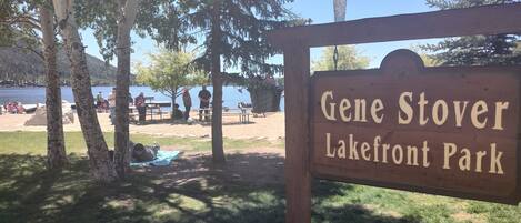 Señal de Gene Stover Lakefront Park con mesas de picnic y gente al fondo.