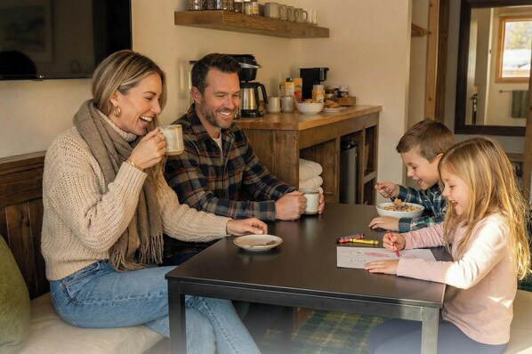 Eine vierköpfige Familie sitzt an einem Tisch mit einer Kaffeemaschine, Schüsseln und einem Teller Essen.