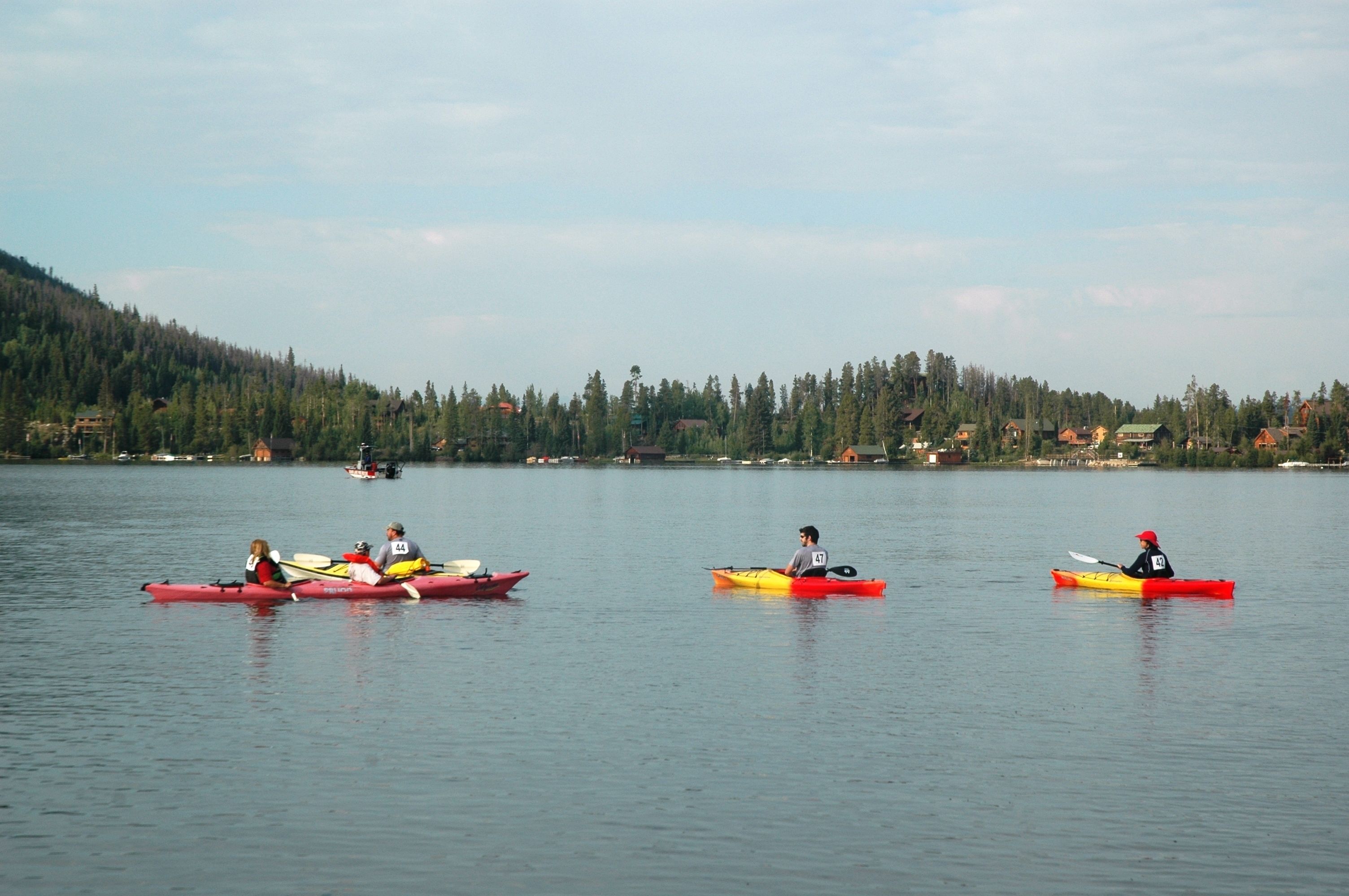 A calm lake with kayaks and canoes, surrounded by forested hills and distant buildings.