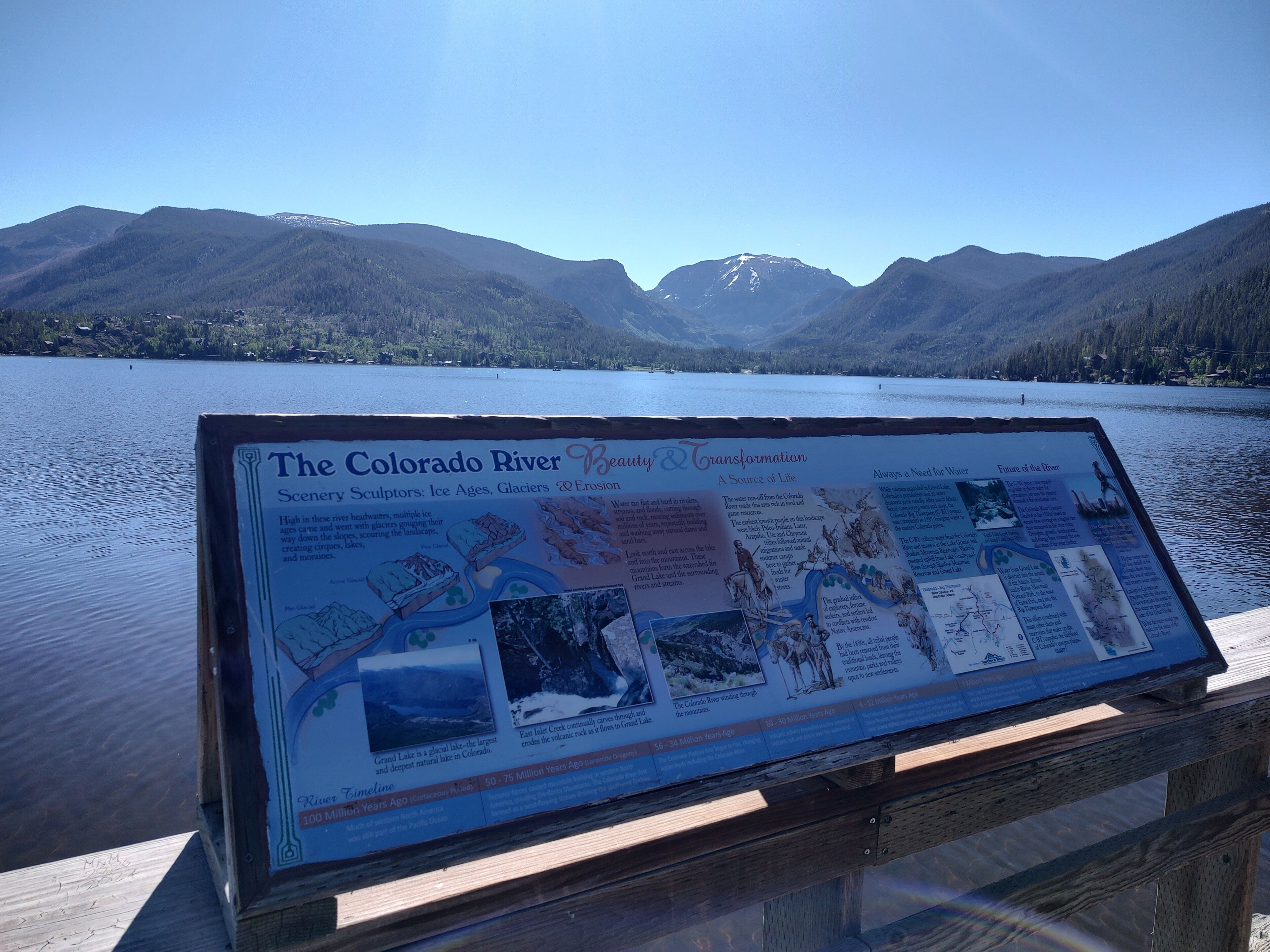 An informational signboard about the Colorado River's history and transformation, placed on a wooden dock by a lake with mountains in the background.