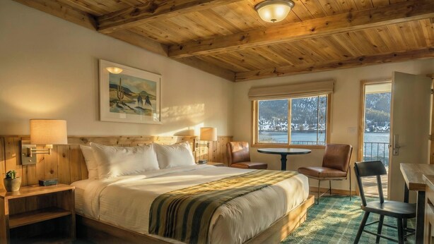 A cozy room with a wooden ceiling, a bed with a green-striped blanket, a painting, a window with a view of a snowy landscape, and a small round table with chairs. - Western Riviera Lakeside Lodging (Grand Lake)