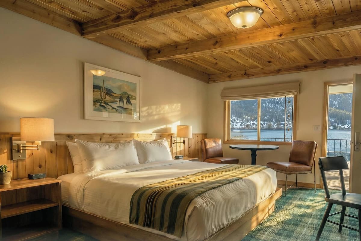 Una habitación acogedora con techo de madera, una cama con manta a rayas verdes, un cuadro, una ventana con vistas a un paisaje nevado y una pequeña mesa redonda con sillas.