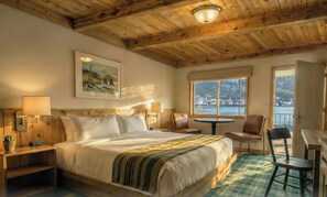 Ein gemütliches Zimmer mit Holzbalkendecke, einem Bett mit grün gestreifter Decke, einem Gemälde, einem Fenster mit Blick auf eine verschneite Landschaft und einem kleinen runden Tisch mit Stühlen.