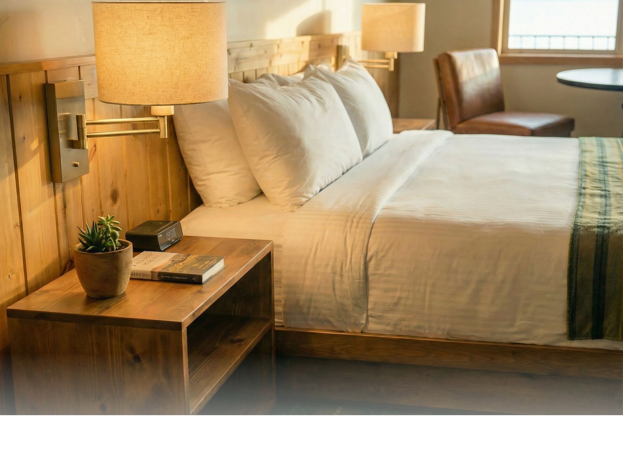 A hotel room with a bed, bedside tables, lamps, a potted plant, and a small table with a book.