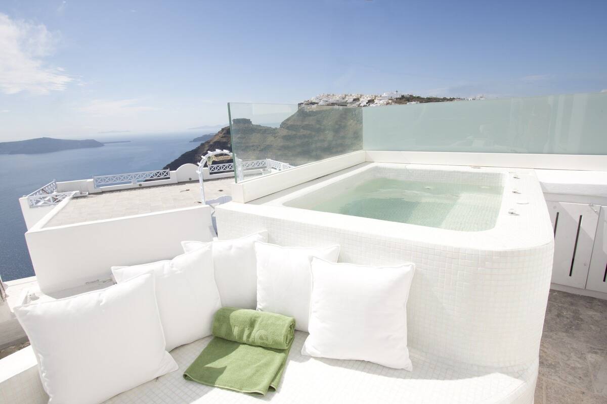 A rooftop terrace with a hot tub, white furniture, and a view of the sea and cliffs.