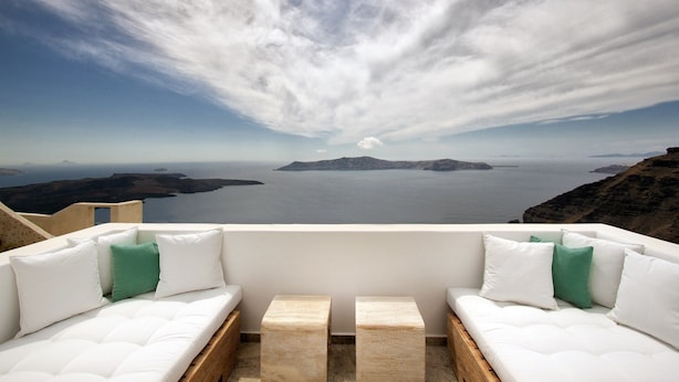 Un balcón con asientos blancos acolchados y vistas a un mar tranquilo y unas islas lejanas.