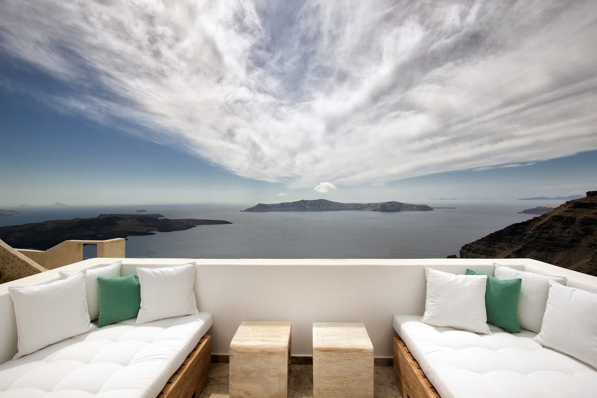 A balcony with white cushioned seating and a view of a calm sea and distant islands.